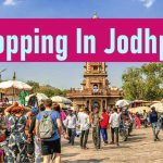 Busy open-air market scene in Jodhpur, India, featuring crowds of shoppers, street stalls, and the historic Ghanta Ghar clock tower in the background under a bright blue sky, overlaid with a pink banner reading "Shopping In Jodhpur."