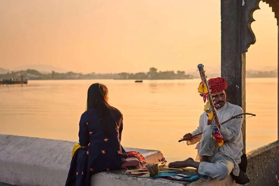 Udaipur, Rajasthan - Woman and Man in Traditional Clothing Sitting on Wall on Lakeshore and Playing Musical Instrument