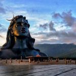 The massive black Adiyogi Shiva statue with a crescent moon on its head stands before green mountains under a dramatic, cloudy sunset sky.