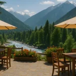 Outdoor cafe in Manali with wooden tables, umbrellas, river view, pine trees, and snow-covered Himalayan mountains in the background on a clear sunny day.